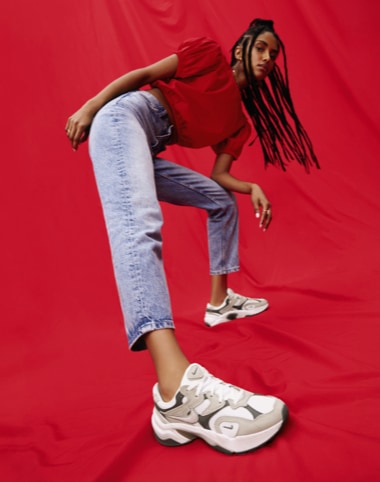 woman in jeans and red shirt leaned over wearing nude and white nike y2k sneakers on red background