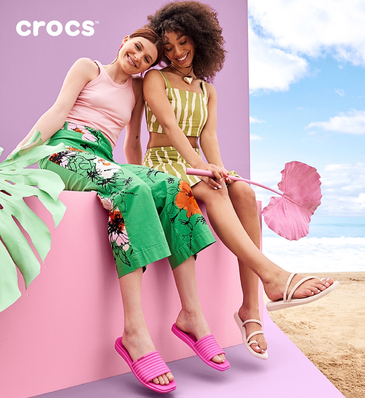 two women sitting down wearing crocs holding giant leaves